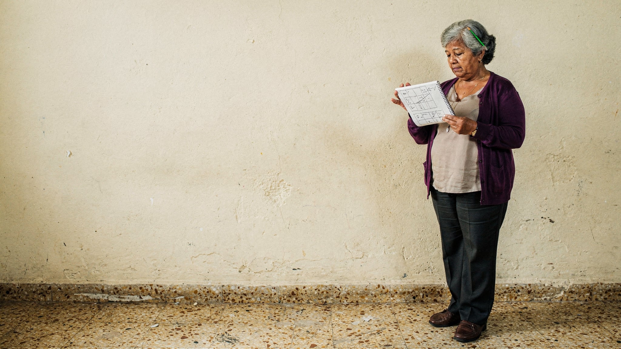 A lottery-shop owner in a violet cardigan holds an open notebook of hand-drawn workflow diagrams against a plain cream wall